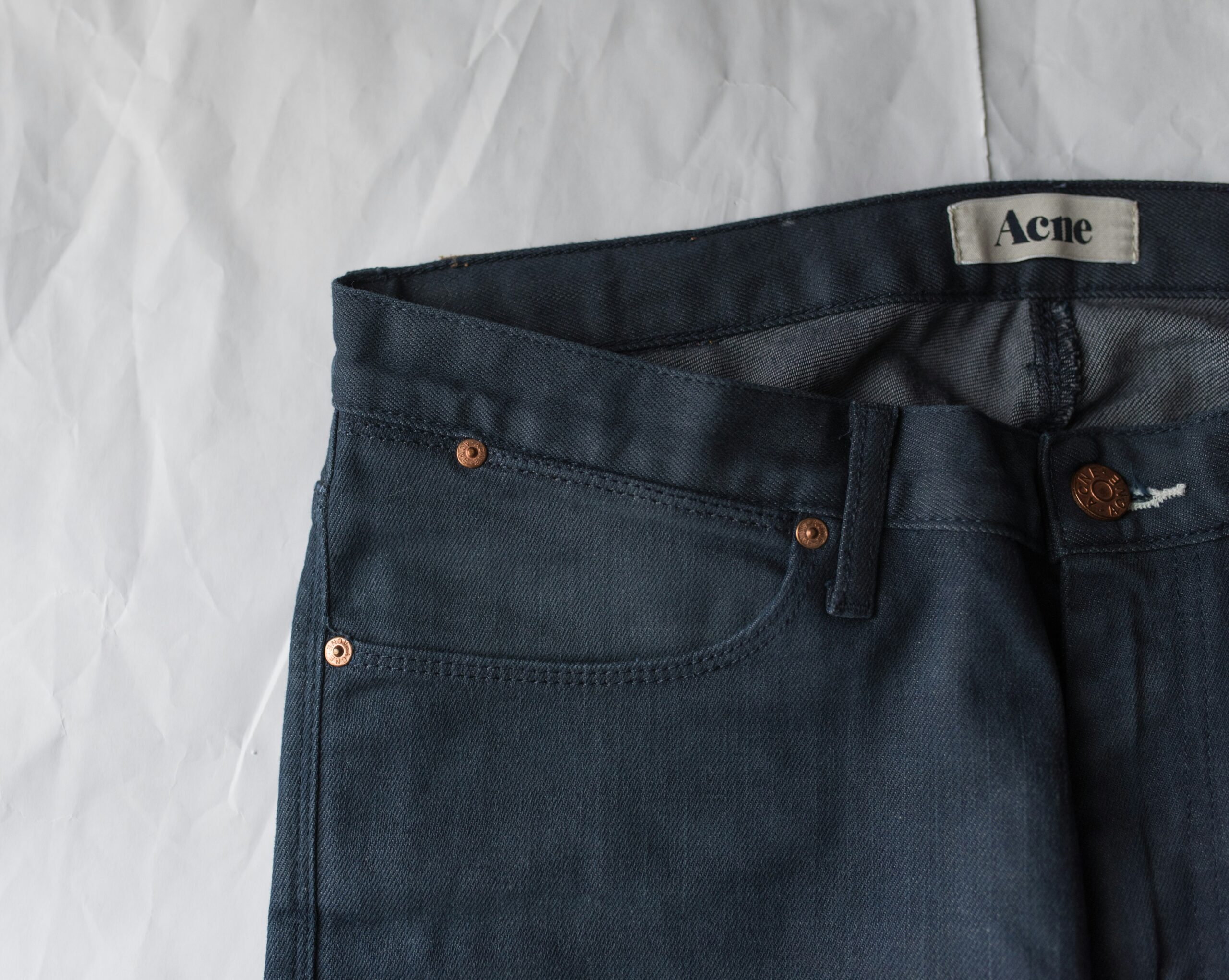 A close-up, top-down view of the waist and front pocket area of a pair of dark blue denim jeans. An "Acne" brand tag is visible on the inner waistband.