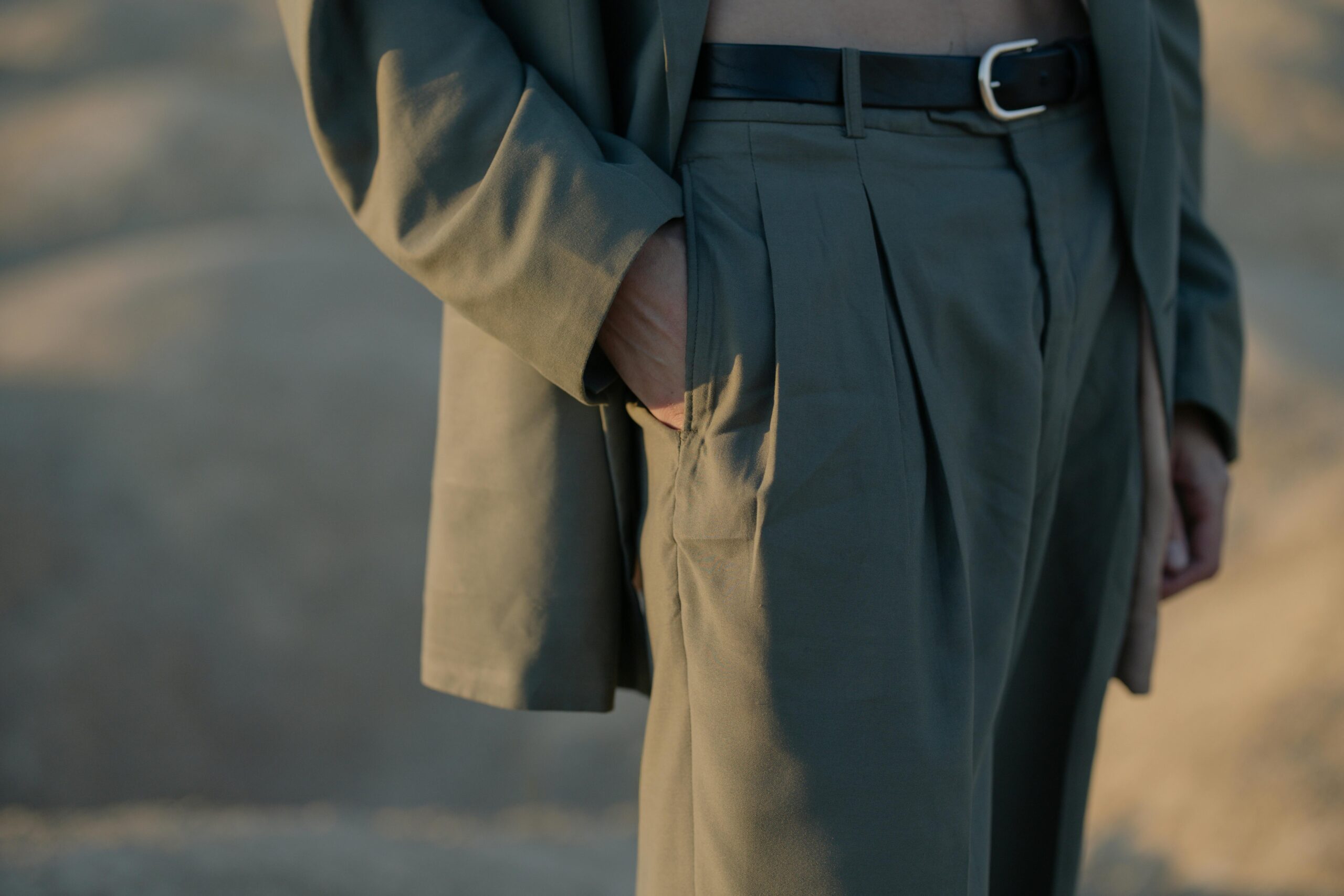 A close-up shot focusing on the side of a person's torso and hip. They are wearing an olive green blazer and matching pleated trousers with a black leather belt. One hand is tucked into the trouser pocket.