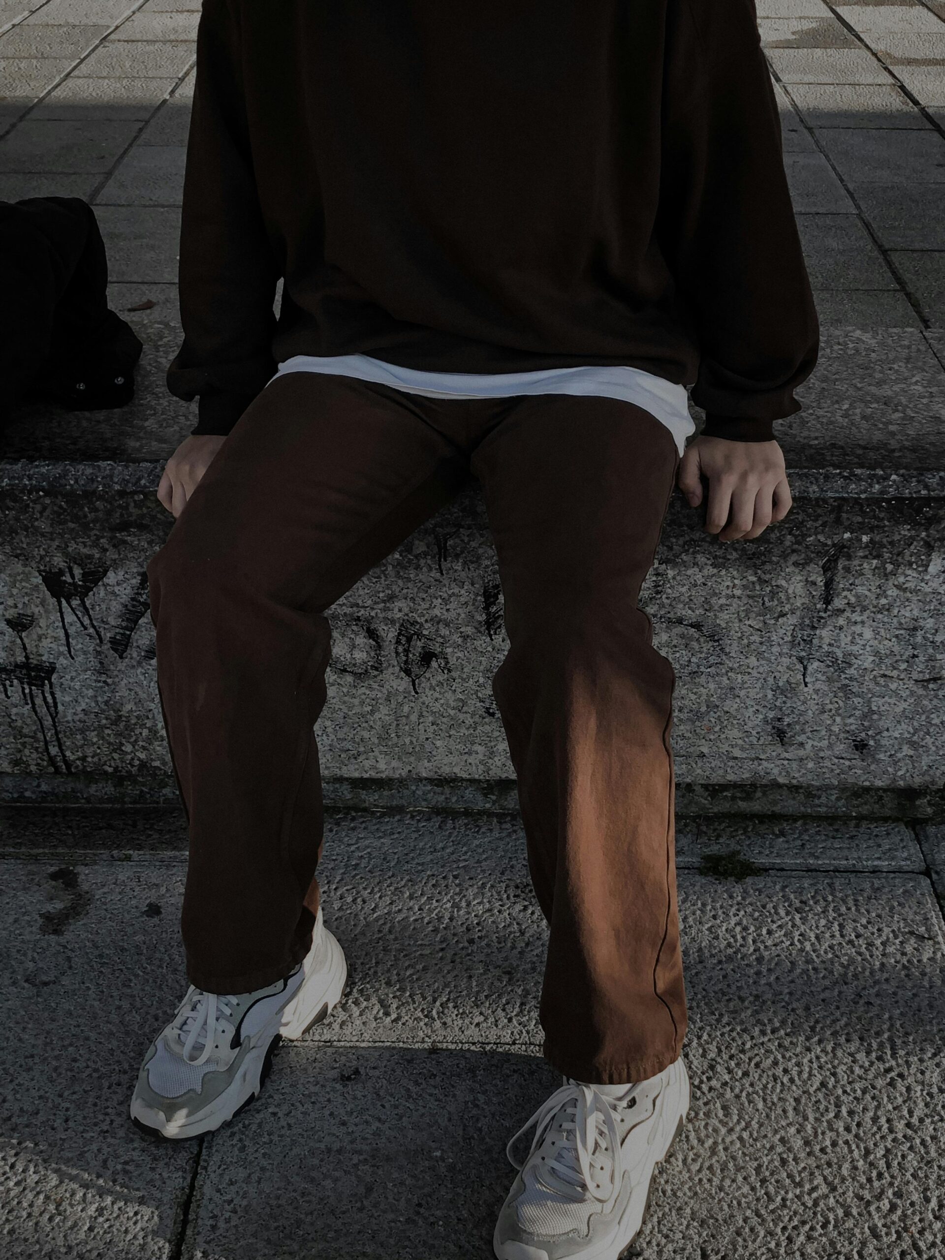 A photograph from the chest down of a person sitting on a stone ledge. They are wearing a dark brown oversized sweatshirt over a white t-shirt, brown trousers, and white chunky sneakers.